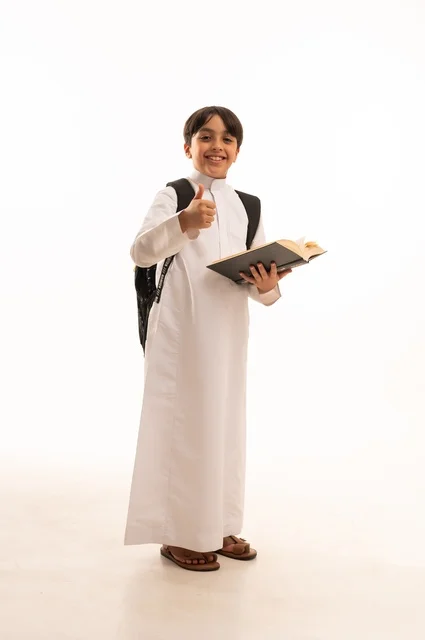 رفع اصبع الابهام بايماءات الرضى والقبول، الاجتهاد للوصول إلى النجاح، المناهج العلمية التعليمية، مفهوم العودة إلى المدارس، بورتريه لطالب عربي خليجي سعودي يرتدي الثوب التقليدي يحمل حقيبة الظهر المدرسية ويقوم بقراءة كتاب، صورة لكامل الجسم، خلفية بيضاء