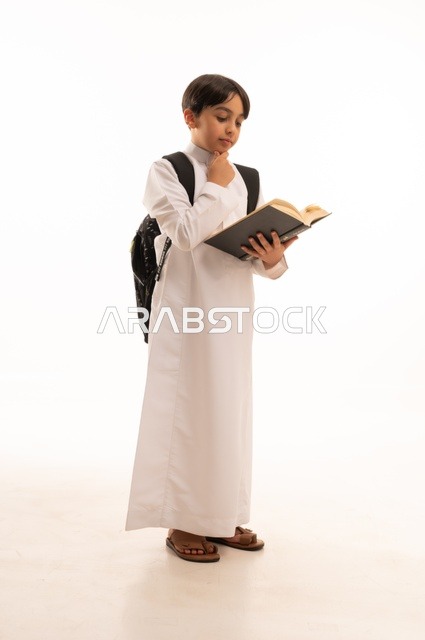 مفهوم العودة إلى المدارس، المناهج العلمية التعليمية، بورتريه لطالب عربي خليجي سعودي يرتدي الثوب التقليدي يحمل حقيبة الظهر المدرسية يضع يده على ذقنه بايماءات التساؤل والاستغراب ويقوم بقراءة كتاب، صورة لكامل الجسم، خلفية بيضاء