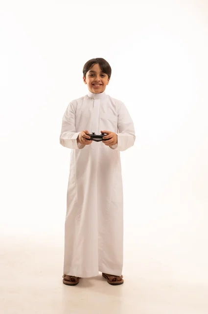 Having fun playing video games, portrait of a Saudi Arabian boy wearing traditional clothing holding a joystick, gestures of enjoyment and integration, using a modern and advanced technological device, white background
