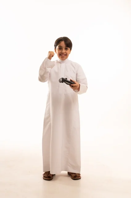 Raising a fist with a victory expression, having a good time playing video games, a portrait of a Saudi Arabian Gulf boy wearing traditional clothing holding a joystick, gestures of enjoyment and integration, using a modern and advanced technological device, white background