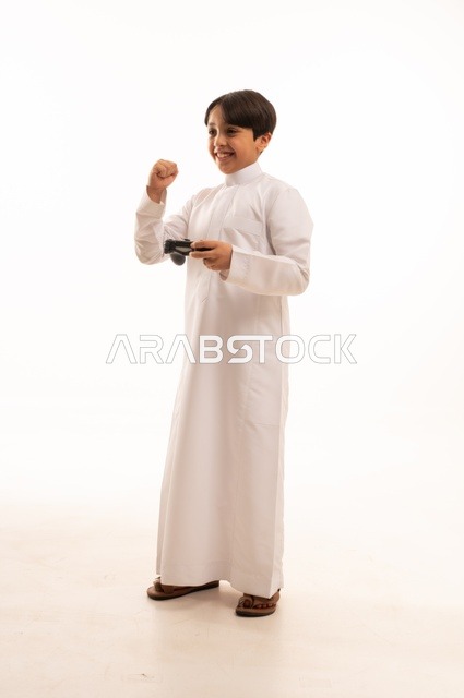 رفع قبضة اليد بتعابير الفوز، قضاء وقت ممتع في ألعاب الفيديو الترفيهيه، بورتريه لولد عربي خليجي سعودي يرتدي الثوب التقليدي يحمل بيده عصا التحكم، إيماءات الاستمتاع والاندماج، استخدام جهاز تكنولوجي تقني حديث ومتطور، خلفية بيضاء