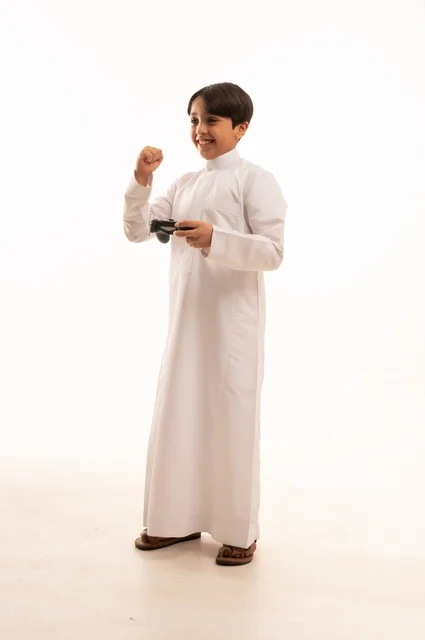 رفع قبضة اليد بتعابير الفوز، قضاء وقت ممتع في ألعاب الفيديو الترفيهيه، بورتريه لولد عربي خليجي سعودي يرتدي الثوب التقليدي يحمل بيده عصا التحكم، إيماءات الاستمتاع والاندماج، استخدام جهاز تكنولوجي تقني حديث ومتطور، خلفية بيضاء