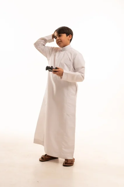 استخدام جهاز تكنولوجي تقني حديث ومتطور، قضاء وقت ممتع في ألعاب الفيديو الترفيهيه، بورتريه لولد عربي خليجي سعودي يرتدي الثوب التقليدي يحمل بيده عصا التحكم ويضع يده على رأسه بإيماءات الخسارة والهزيمة، الجسم بالطول الكامل، خلفية بيضاء