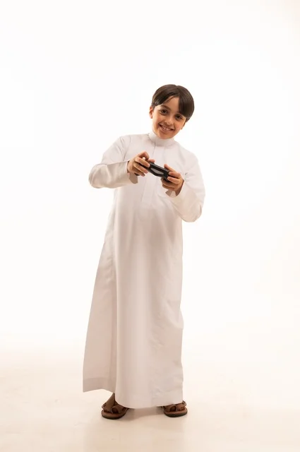 قضاء وقت ممتع في ألعاب الفيديو الترفيهيه، بورتريه لولد عربي خليجي سعودي يرتدي الثوب التقليدي يحمل بيده عصا التحكم، إيماءات الاستمتاع والاندماج، استخدام جهاز تكنولوجي تقني حديث ومتطور، الجسم بالطول الكامل، خلفية بيضاء