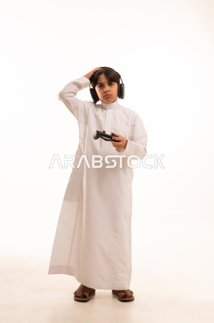 استخدام جهاز تكنولوجي تقني حديث ومتطور، قضاء وقت ممتع في ألعاب الفيديو الترفيهيه، بورتريه لولد عربي خليجي سعودي يرتدي الثوب التقليدي يحمل بيده عصا التحكم ويضع يده على رأسه بإيماءات الخسارة والهزيمة، الجسم بالطول الكامل، خلفية بيضاء