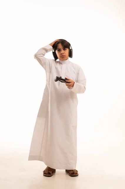 استخدام جهاز تكنولوجي تقني حديث ومتطور، قضاء وقت ممتع في ألعاب الفيديو الترفيهيه، بورتريه لولد عربي خليجي سعودي يرتدي الثوب التقليدي يحمل بيده عصا التحكم ويضع يده على رأسه بإيماءات الخسارة والهزيمة، الجسم بالطول الكامل، خلفية بيضاء