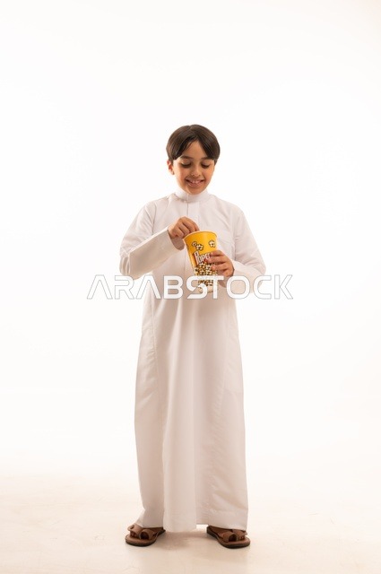 الهروب من الروتين اليومي والقيام بنشاط ترفيهي مسلي، الاستمتاع بمشاهدة الأفلام المفضلة،  تناول الفوشار اللذيذ والمقرمش، بورتريه لولد عربي خليجي سعودي يرتدي الثوب التقليدي يمسك بيده كيس من الفوشار، الجسم بالطول الكامل، خلفية بيضاء