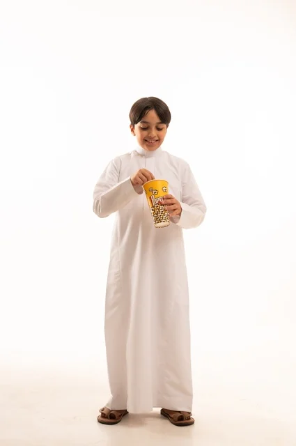 الهروب من الروتين اليومي والقيام بنشاط ترفيهي مسلي، الاستمتاع بمشاهدة الأفلام المفضلة،  تناول الفوشار اللذيذ والمقرمش، بورتريه لولد عربي خليجي سعودي يرتدي الثوب التقليدي يمسك بيده كيس من الفوشار، الجسم بالطول الكامل، خلفية بيضاء