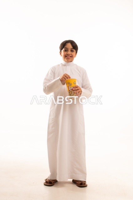الهروب من الروتين اليومي والقيام بنشاط ترفيهي مسلي، الاستمتاع بمشاهدة الأفلام المفضلة،  تناول الفوشار اللذيذ والمقرمش، بورتريه لولد عربي خليجي سعودي يرتدي الثوب التقليدي يمسك بيده كيس من الفوشار، الجسم بالطول الكامل، خلفية بيضاء