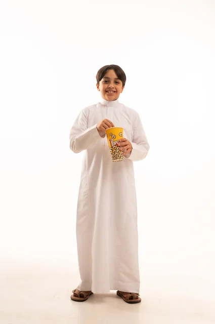 Escape from daily routine and do an entertaining recreational activity, enjoy watching your favorite movies, eat delicious and crunchy popcorn, portrait of a Saudi Arabian Gulf boy wearing traditional dress holding a bag of popcorn, full body, white background
