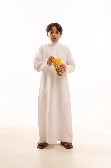 الهروب من الروتين اليومي والقيام بنشاط ترفيهي مسلي، الاستمتاع بمشاهدة الأفلام المفضلة،  تناول الفوشار اللذيذ والمقرمش، بورتريه لولد عربي خليجي سعودي يرتدي الثوب التقليدي يمسك بيده كيس من الفوشار، الجسم بالطول الكامل، خلفية بيضاء
