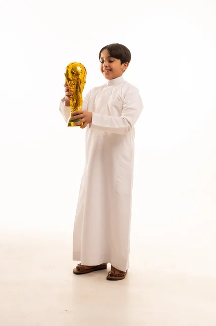 تعابير الفرح والسرور والحماس للمنافسة، مفهوم الربح والفوز، تشجيع الفريق السعودي، التتويج بالبطولة والحصول على المركز الأول، بورتريه لولد عربي خليجي سعودي يرتدي الثوب التقليدي يحمل بيده كأس كرة قدم ذهبي, صورة الجسم بالطول الكامل
, خلفية بيضاء