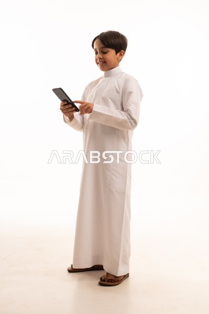 التواصل وتبادل الأحاديث مع الأهل والأصدقاء عبر الهاتف المحمول، بورتريه لولد عربي خليجي سعودي يرتدي الثوب التقليدي يقوم بالتحدث عبر الجوال بايماءات السعادة والسرور، استخدام جهاز تقني حديث ومتطور، الجسم بالطول الكامل, خلفية بيضاء
