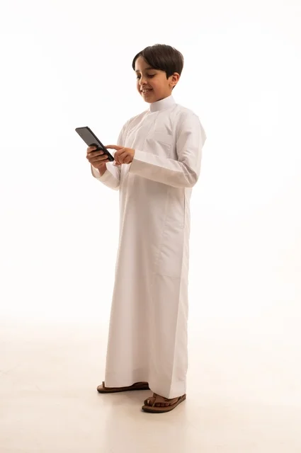 Communicating and exchanging conversations with family and friends via mobile phone, portrait of a Saudi Arabian Gulf boy wearing traditional dress talking on the mobile phone with gestures of happiness and pleasure, using a modern and advanced technical device, full body, white background