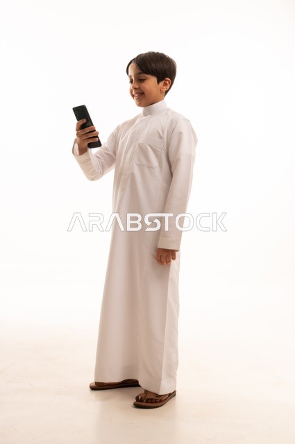 Communicating and exchanging conversations with family and friends via mobile phone, portrait of a Saudi Arabian Gulf boy wearing traditional dress talking on the mobile phone with gestures of happiness and pleasure, using a modern and advanced technical device, full body, white background