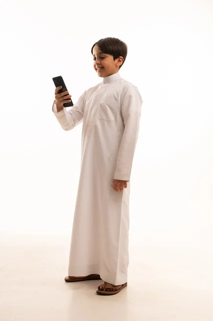 التواصل وتبادل الأحاديث مع الأهل والأصدقاء عبر الهاتف المحمول، بورتريه لولد عربي خليجي سعودي يرتدي الثوب التقليدي يقوم بالتحدث عبر الجوال بايماءات السعادة والسرور، استخدام جهاز تقني حديث ومتطور، الجسم بالطول الكامل, خلفية بيضاء