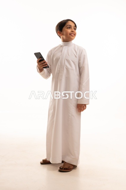 التواصل وتبادل الأحاديث مع الأهل والأصدقاء عبر الهاتف المحمول، بورتريه لولد عربي خليجي سعودي يرتدي الثوب التقليدي يقوم بالتحدث عبر الجوال بايماءات السعادة والسرور، استخدام جهاز تقني حديث ومتطور، الجسم بالطول الكامل, خلفية بيضاء
