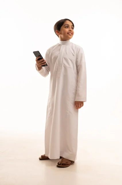 التواصل وتبادل الأحاديث مع الأهل والأصدقاء عبر الهاتف المحمول، بورتريه لولد عربي خليجي سعودي يرتدي الثوب التقليدي يقوم بالتحدث عبر الجوال بايماءات السعادة والسرور، استخدام جهاز تقني حديث ومتطور، الجسم بالطول الكامل, خلفية بيضاء