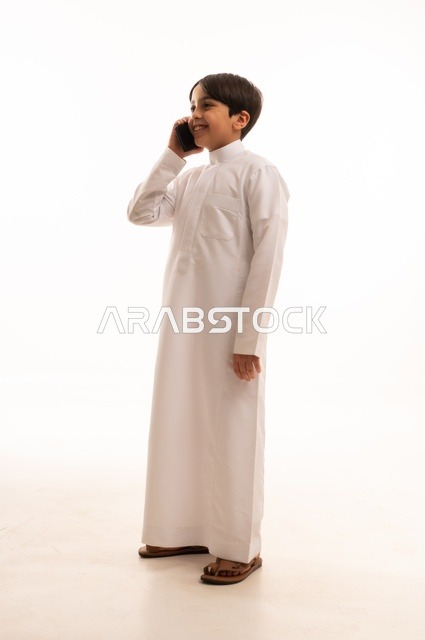 Making a phone call on a mobile phone, portrait of a Saudi Arabian Gulf boy wearing traditional dress looking at a place with gestures of happiness and pleasure, hearing good news, using a modern and advanced technical device, full body, white background