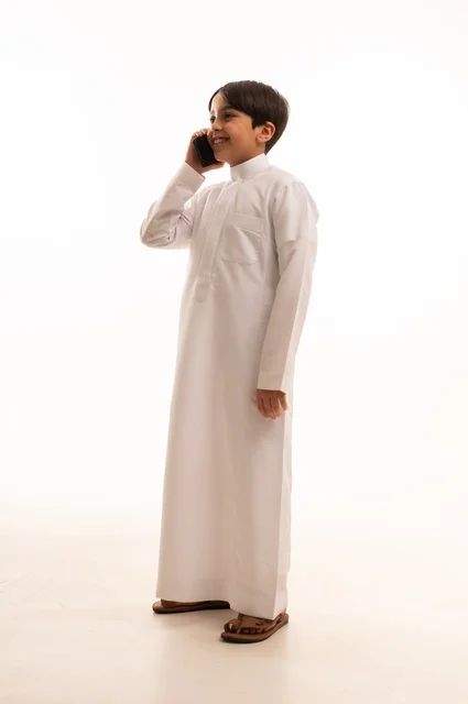Making a phone call on a mobile phone, portrait of a Saudi Arabian Gulf boy wearing traditional dress looking at a place with gestures of happiness and pleasure, hearing good news, using a modern and advanced technical device, full body, white background