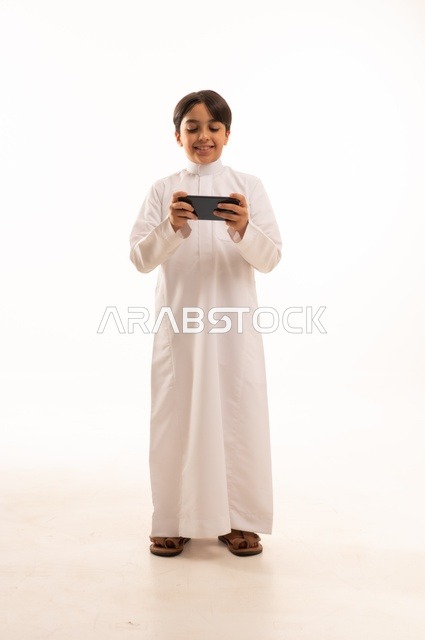 التواصل وتبادل الأحاديث مع الأهل والأصدقاء عبر الهاتف المحمول، بورتريه لولد عربي خليجي سعودي يرتدي الثوب التقليدي يقوم بالتحدث عبر الجوال بايماءات السعادة والسرور، استخدام جهاز تقني حديث ومتطور، الجسم بالطول الكامل, خلفية بيضاء
