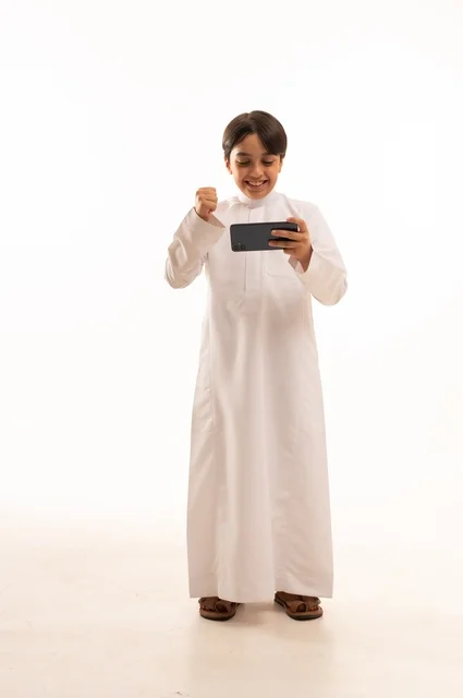 رفع قبضة اليد بتعابير الفوز والانتصار، بورتريه لولد عربي خليجي سعودي يرتدي الثوب التقليدي يقوم بمشاهدة الافلام والمباراة عبر الهاتف المحمول،  قضاء وقت ممتع في العطلة، استخدام جهاز تقني حديث ومتطور، الجسم بالطول الكامل, خلفية بيضاء