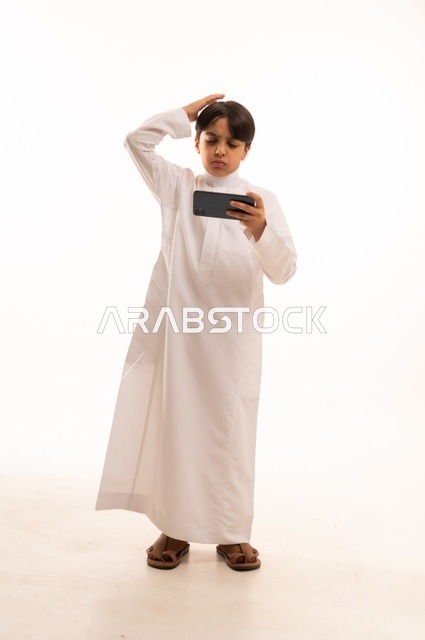 وضع اليد على الراس بايماءات الخسارة، بورتريه لولد عربي خليجي سعودي يرتدي الثوب التقليدي يقوم بمشاهدة الافلام والمباراة عبر الهاتف المحمول،  قضاء وقت ممتع في العطلة، استخدام جهاز تقني حديث ومتطور، الجسم بالطول الكامل, خلفية بيضاء