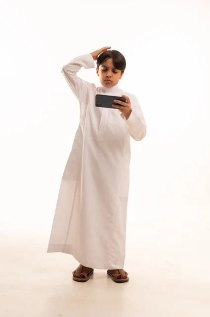 وضع اليد على الراس بايماءات الخسارة، بورتريه لولد عربي خليجي سعودي يرتدي الثوب التقليدي يقوم بمشاهدة الافلام والمباراة عبر الهاتف المحمول،  قضاء وقت ممتع في العطلة، استخدام جهاز تقني حديث ومتطور، الجسم بالطول الكامل, خلفية بيضاء