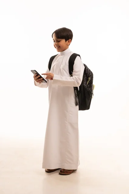 مفهوم العودة إلى المدارس، المناهج العلمية التعليمية، بورتريه لولد عربي خليجي سعودي يرتدي الثوب التقليدي وحقيبة الظهر يحمل بيده الهاتف المحمول، التواصل وتبادل الاحاديث مع الاهل والاصدقاء، استخدام جهاز تقني حديث ومتطور، الجسم بالطول الكامل, خلفية بيضاء