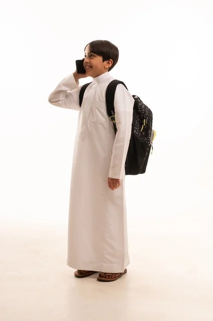 Educational science curricula, back to school concept, portrait of a Saudi Arabian boy wearing traditional clothing and a backpack holding a mobile phone, making a phone call, using a modern and advanced technical device, full body, white background