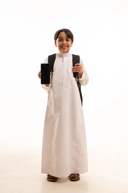 المناهج العلمية التعليمية، مفهوم العودة إلى المدارس،  بورتريه لولد عربي خليجي سعودي يرتدي الثوب التقليدي وحقيبة الظهر يحمل بيده الهاتف المحمول، عرض شاشة سوداء فارغة عبر الجوال، استخدام جهاز تقني حديث ومتطور، الجسم بالطول الكامل, خلفية بيضاء