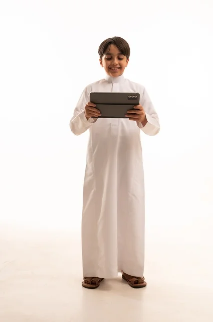 The concept of e-learning, following lessons remotely via the Internet, educational scientific curricula, a portrait of a Saudi Arabian Gulf boy wearing traditional clothing and a backpack, holding a tablet in his hand, using a modern and advanced technical device, full-length body, white background
