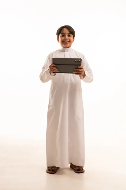 مفهوم التعليم الالكتروني، متابعة الدروس عن بعد عن طريق الانترنت، المناهج العلمية التعليمية،  بورتريه لولد عربي خليجي سعودي يرتدي الثوب التقليدي وحقيبة الظهر يحمل بيده الجهاز اللوحي، استخدام جهاز تقني حديث ومتطور، الجسم بالطول الكامل, خلفية بيضاء