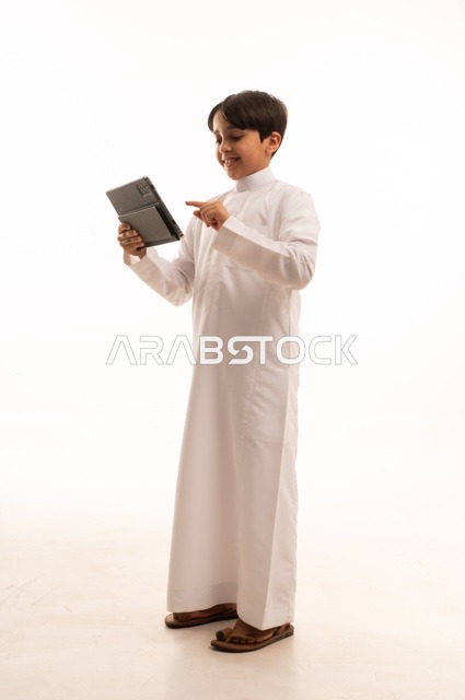 مفهوم التعليم الالكتروني، متابعة الدروس عن بعد عن طريق الانترنت، المناهج العلمية التعليمية،  بورتريه لولد عربي خليجي سعودي يرتدي الثوب التقليدي وحقيبة الظهر يحمل بيده الجهاز اللوحي، استخدام جهاز تقني حديث ومتطور، الجسم بالطول الكامل, خلفية بيضاء