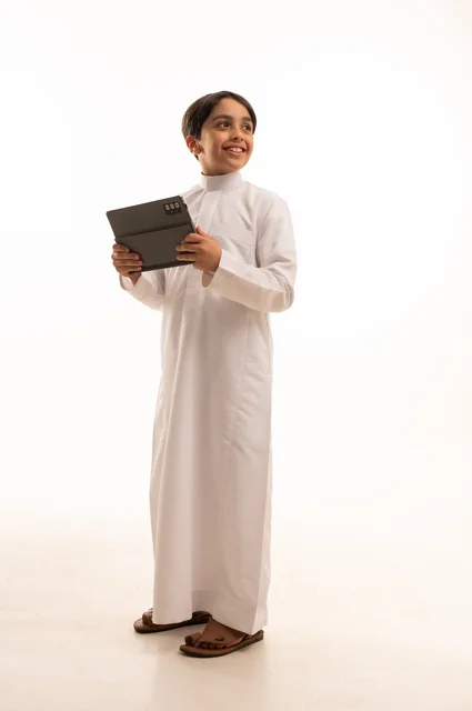 مفهوم التعليم الالكتروني، متابعة الدروس عن بعد عن طريق الانترنت، المناهج العلمية التعليمية،  بورتريه لولد عربي خليجي سعودي يرتدي الثوب التقليدي وحقيبة الظهر يحمل بيده الجهاز اللوحي، استخدام جهاز تقني حديث ومتطور، الجسم بالطول الكامل, خلفية بيضاء