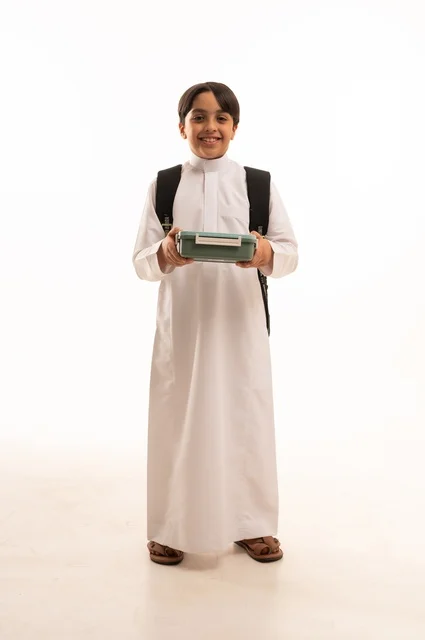 The importance of eating healthy and balanced food to maintain a healthy body and mind. Portrait of a Saudi Arabian Gulf student wearing traditional thobe and backpack holding a lunch box, snack and healthy meal for school break. Back to school season in Saudi Arabia. Full body, white background.