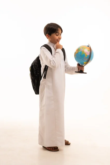الاهتمام بدراسة العلوم الجغرافية، توسيع الآفاق وفهم التنوع الثقافي والطبيعي، أهمية التعليم في تطوير المجتمع السعودي، بورتريه لطالب عربي خليجي سعودي يرتدي الثوب التقليدي وحقيبة الظهر يحمل بيده مجسم الكرة الأرضية، الجسم بالطول الكامل، خلفية بيضاء