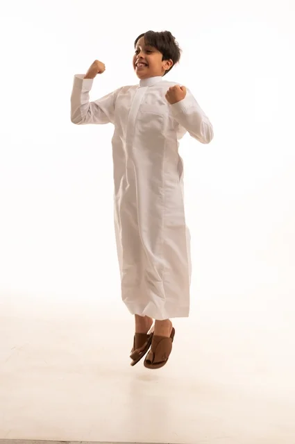 مفهوم اللهو واللعب، القفز عالياً بايماءات السعادة والسرور، بورتريه لولد عربي خليجي سعودي يرتدي الثوب التقليدي يقفز في الجو، قضاء وقت ممتع ومسلي، الجسم بالطول الكامل، خلفية بيضاء