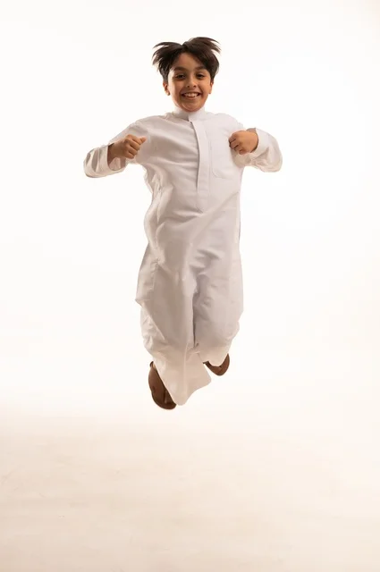 مفهوم اللهو واللعب، القفز عالياً بايماءات السعادة والسرور، بورتريه لولد عربي خليجي سعودي يرتدي الثوب التقليدي يقفز في الجو، قضاء وقت ممتع ومسلي، الجسم بالطول الكامل، خلفية بيضاء