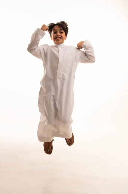 مفهوم اللهو واللعب، القفز عالياً بايماءات السعادة والسرور، بورتريه لولد عربي خليجي سعودي يرتدي الثوب التقليدي يقفز في الجو، قضاء وقت ممتع ومسلي، الجسم بالطول الكامل، خلفية بيضاء