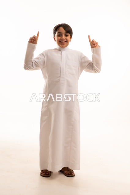 حركات تدل على هذا المكان (هنا)، ايماءات تحديد موقع معين، بورتريه لولد عربي خليجي سعودي يرتدي الثوب التقليدي ويشير باصبع السبابة الى شيء ما، صورة الجسم بالطول الكامل، خلفية بيضاء