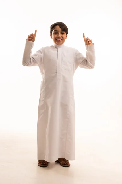 حركات تدل على هذا المكان (هنا)، ايماءات تحديد موقع معين، بورتريه لولد عربي خليجي سعودي يرتدي الثوب التقليدي ويشير باصبع السبابة الى شيء ما، صورة الجسم بالطول الكامل، خلفية بيضاء