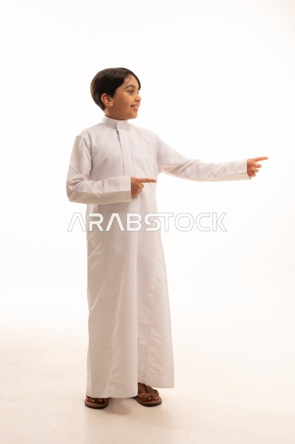 ايماءات تحديد موقع معين، حركات تدل على هذا المكان (هنا)، بورتريه لولد عربي خليجي سعودي يرتدي الثوب التقليدي ويشير باصبع السبابة الى شيء ما، صورة الجسم بالطول الكامل، خلفية بيضاء