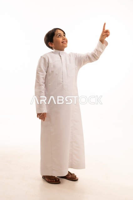 حركات تدل على هذا المكان (هنا)، ايماءات تحديد موقع معين، بورتريه لولد عربي خليجي سعودي يرتدي الثوب التقليدي ويشير باصبع السبابة الى شيء ما، صورة الجسم بالطول الكامل، خلفية بيضاء