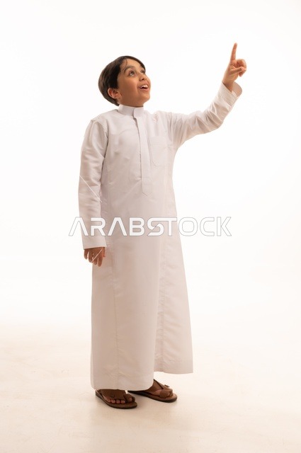 حركات تدل على هذا المكان (هنا)، ايماءات تحديد موقع معين، بورتريه لولد عربي خليجي سعودي يرتدي الثوب التقليدي ويشير باصبع السبابة الى شيء ما، صورة الجسم بالطول الكامل، خلفية بيضاء