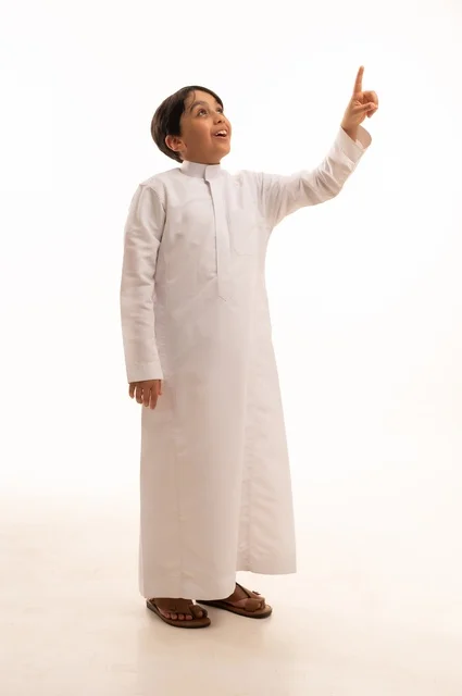 حركات تدل على هذا المكان (هنا)، ايماءات تحديد موقع معين، بورتريه لولد عربي خليجي سعودي يرتدي الثوب التقليدي ويشير باصبع السبابة الى شيء ما، صورة الجسم بالطول الكامل، خلفية بيضاء