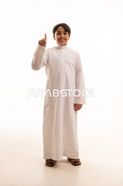 حركات تدل على هذا المكان (هنا)، ايماءات تحديد موقع معين، بورتريه لولد عربي خليجي سعودي يرتدي الثوب التقليدي ويشير باصبع السبابة الى شيء ما، صورة الجسم بالطول الكامل، خلفية بيضاء