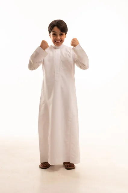 Achievement and goal attainment, interest in elegance and outward appearance, concept of self-confidence, portrait of a Saudi Arabian Gulf boy wearing traditional dress and raising his fists in the air with gestures of victory, triumph and success, full-length body, white background