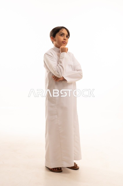 النظر الى شيء ما بايماءات التفكير، الاهتمام بالأناقة والمظهر الخارجي، مفهوم الثقة بالنفس، بورتريه لولد عربي خليجي سعودي يرتدي الثوب التقليدي يضع يده على ذقنه، الجسم بالطول الكامل، خلفية بيضاء