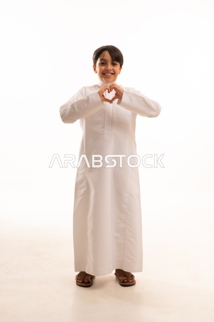 Showing feelings of love and passion, the concept of love and affection, interest in elegance and outward appearance, portrait of a Saudi Arabian Gulf boy wearing traditional dress raising his hand in the shape of a heart with gestures of pleasure and happiness, full body, white background